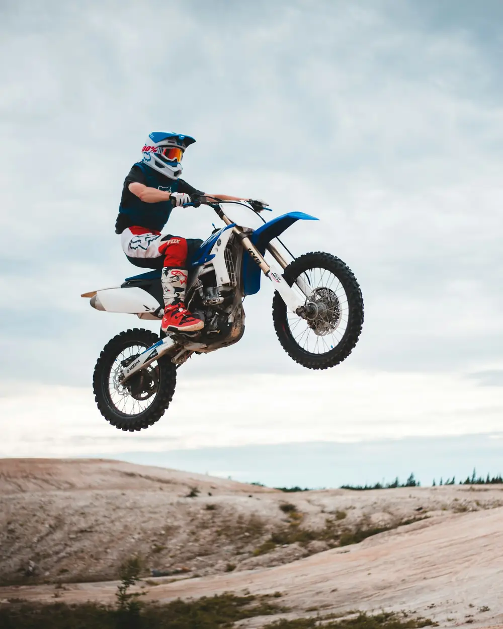 Piloto de motocross em pleno salto com moto azul e branca sobre terreno arenoso